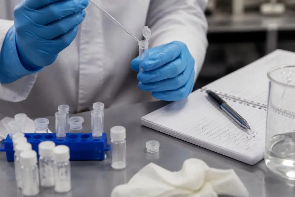 Lab technician in blue gloves uses a pipette to transfer liquid, with vials and a notebook on the bench