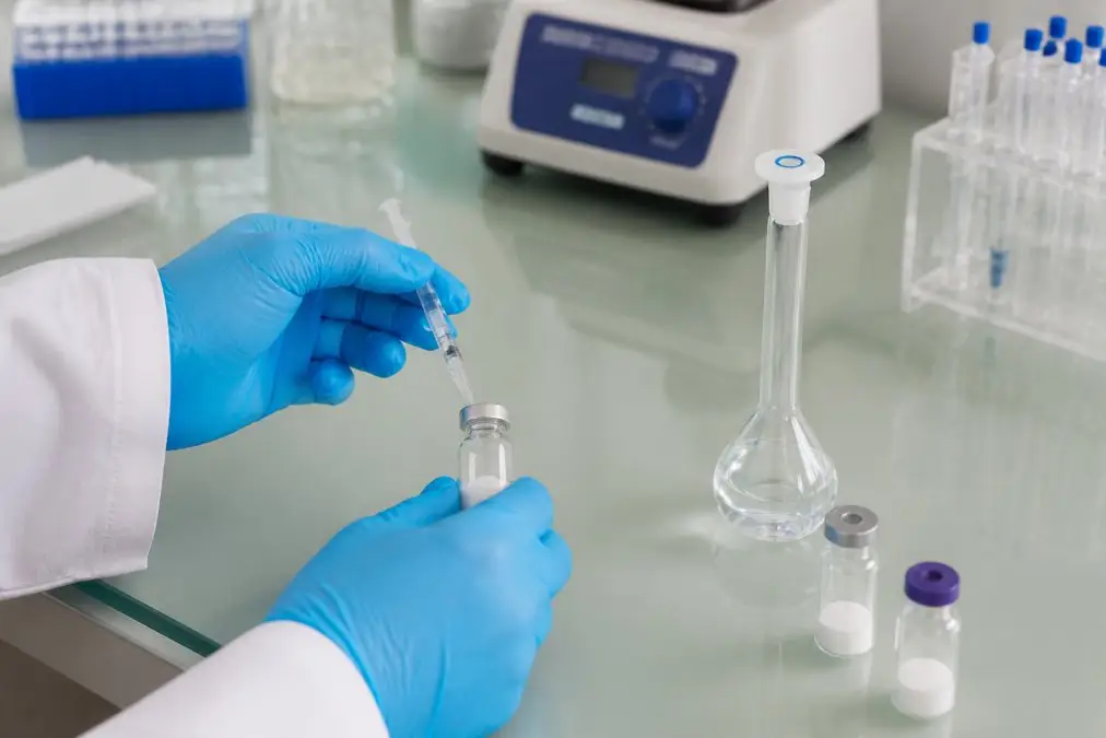 Gloved hands drawing liquid from a vial with a syringe in a laboratory setup on a glass bench.